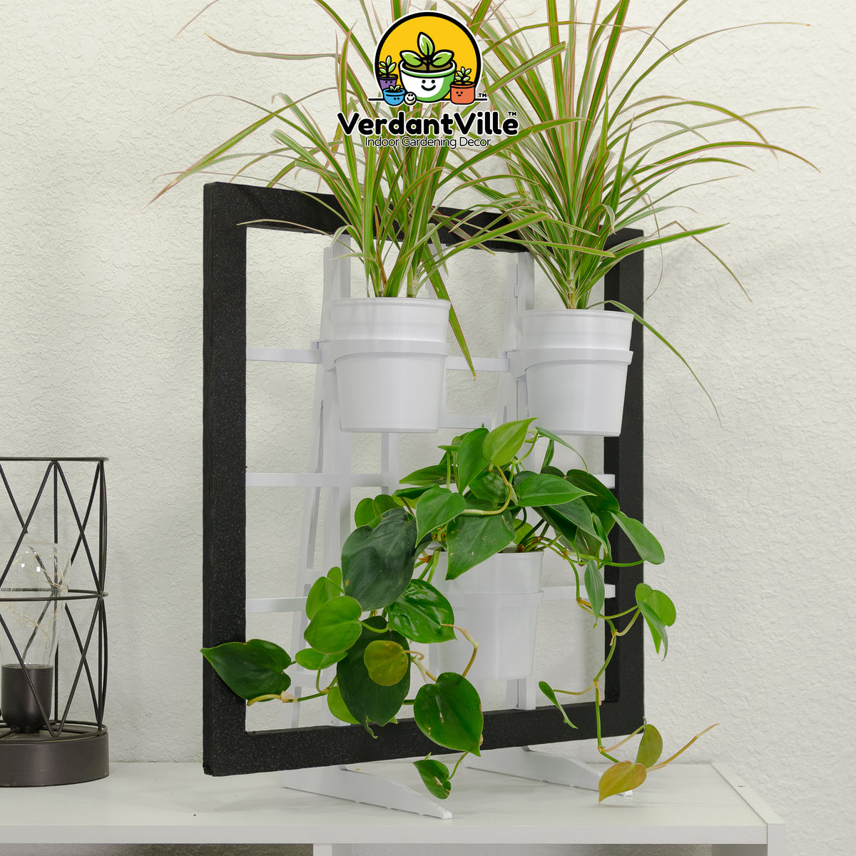 A free-standing plant holder with a black Frame, three white hanging pots, styled with green houseplants against a white textured wall. A VerdantVille logo watermark is visible at the top, and a small black decorative lantern sits on the table to the left.