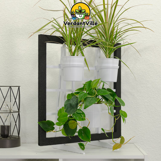 A free-standing plant holder with a black Frame, three white hanging pots, styled with green houseplants against a white textured wall. A VerdantVille logo watermark is visible at the top, and a small black decorative lantern sits on the table to the left.